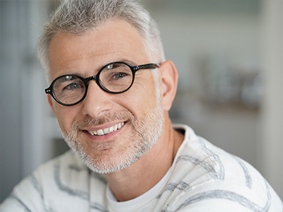 The image shows a man with glasses and a beard, smiling slightly at the camera. He has short hair and appears to be middle-aged. The background is blurred but suggests an indoor setting with a neutral color palette.